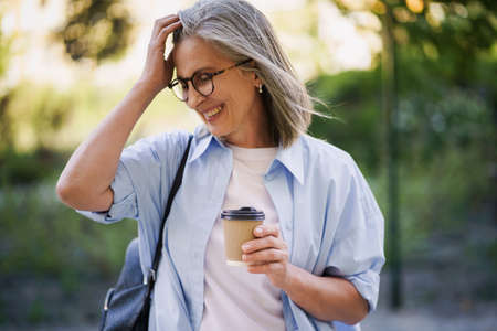 Charming mature woman drinks coffee on the go fixing her hair. Mature woman spend free time traveling in european old town enjoying cup of tea, coffee. Grey haired woman in casual with backpack.の写真素材