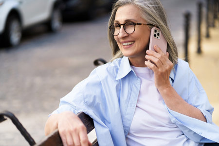 Enjoying summer time mature woman with grey hair talking on the phone holding smartphone sitting on the bench at the streets of old european town. Mature woman answering call outdoors.の写真素材