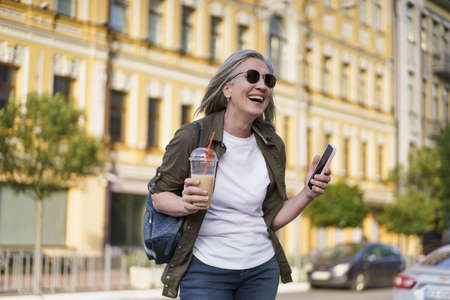 European senior woman with gray hair happy enjoying free time after work or traveling holding phone while having juice on the go using plastic cup in city background. Enjoying life mature woman.の写真素材