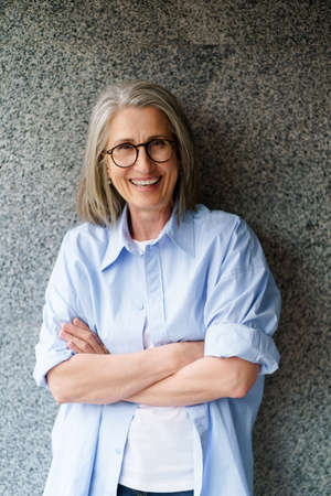 Portrait of charming mature woman with grey hair standing outdoors leaned on marble wall smiling looking at camera with folded arms. Standing outdoors positive gorgeous woman in casual blue shirt.の写真素材