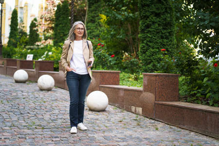 Beautiful mature business woman in casual formal with phone in hand walking on street of old town from work or back to office. Mature woman spend free time traveling in european city.の写真素材