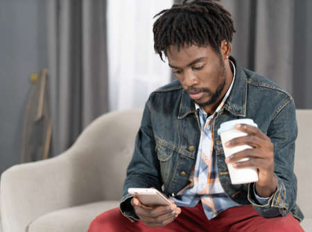 African American young man holding paper or disposable cup with plastic lid and smartphone in hand. Portrait of happy man having present conversation sitting on the sofa. Social media concept.の写真素材