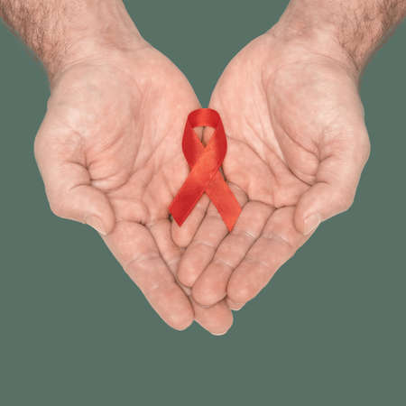 Red awareness ribbon bow on mans helping hands isolated on green background. HIV, AIDS world day. Social life issues concept. Aids charity fund concept. Healthcare and medicine concept.の写真素材
