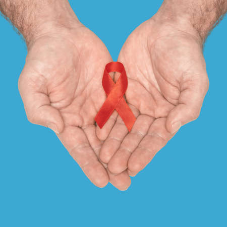 Red awareness ribbon bow on mans helping hands isolated on blue background. HIV, AIDS world day. Social life issues concept. Aids charity fund concept. Healthcare and medicine concept.の写真素材