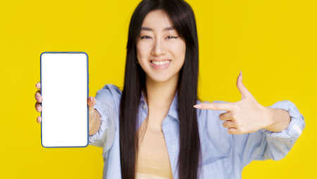 Mobile phone app advertisement. Young asian girl happy holding smartphone pointing on white empty screen and exciting to win isolated on yellow background. Product placement. Selective focus on phone.の写真素材
