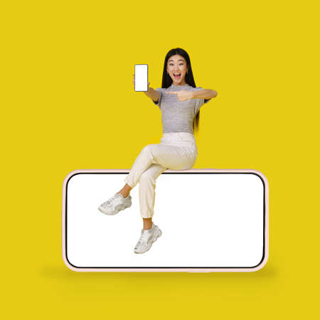 Asian young girl holding smartphone sitting on giant, huge smartphone with black screen isolated on yellow background. Mock up product placement. App Advertisement. Copy space.の写真素材