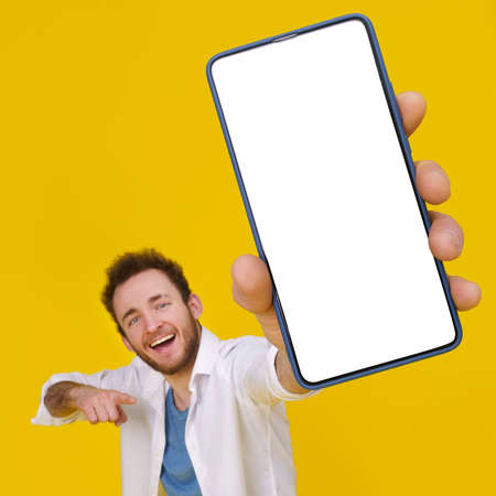 Young happy man holding smartphone showing a white empty screen check this out, cellphone display mock up mobile app advertisement. isolated on yellow background, Product placement.の写真素材