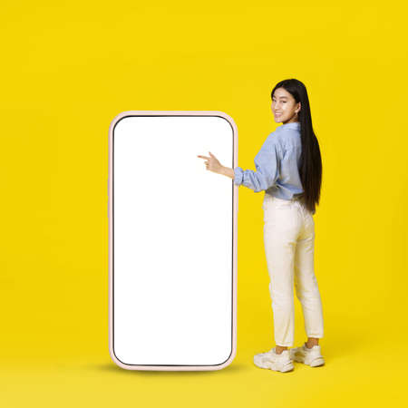Asian girl pointing on giant, huge smartphone with white blank screen standing back happy pointing finger up dressed in casual isolated on yellow background. Free space mock up.の写真素材