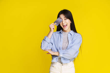 Happy asian girl holding credit, debit card touching her face, eye. Asian girl in blue shirt holding mockup credit card isolated on yellow background. Shopping concept.の写真素材