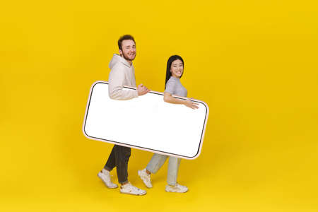 Asian girl and caucasian guy holding huge smartphone showing blank white screen, mobile app advertisement and excited smile on camera isolated on yellow background. Product placement.の写真素材