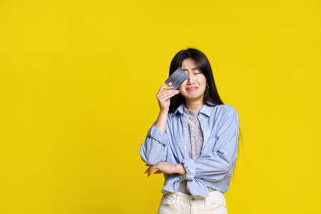 crying unhappy asian girl holding dark credit, debit card touching her eye hiding begging for money. Asian girl in blue shirt with mockup credit card isolated on yellow background. Shopping concept.の写真素材