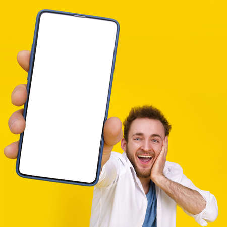 Great offer. Young happy man holding smartphone showing a white empty screen and exciting to win isolated over yellow background, celebrating success. Product placement for mobile app advertisement.の写真素材