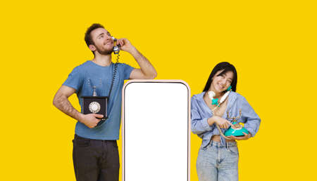 Asian girl and caucasian guy in love talking on vintage phones leaned on huge smartphone with blank screen, looking at each other isolated on yellow background. Product placement.の写真素材