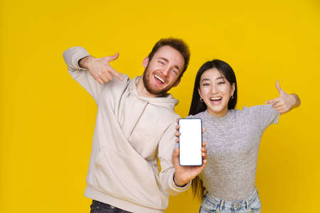 Asian girl and caucasian guy showing thumbs up holding smartphone showing blank white screen, mobile app advertisement and excited smile on camera isolated on yellow background. Product placement.の写真素材