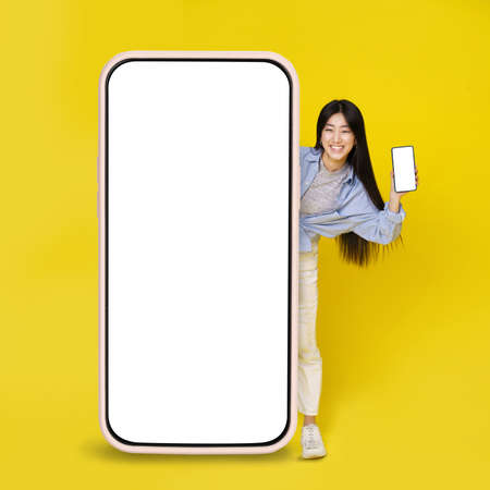 Cute asian young girl excited peeking out big giant vertical cell phone holding smaller one in hand with white blank screen isolated on yellow background. Great offer. Product placement. Copy space.の写真素材