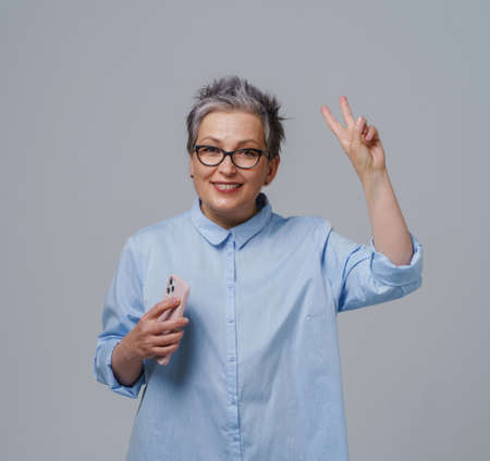 Grey haired gorgeous mature businesswoman with smartphone in hand looking at camera working or shopping online or checking on social media. Pretty woman in blue shirt isolated on white. Toned image.の写真素材