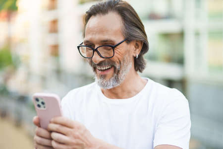 Middle aged grey beard man video calling using smartphone camera standing outdoors with city streets background wearing white shirt. Happy middle aged man on street of european city with mobile phone.の写真素材