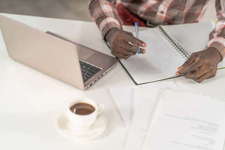 Top view, young african student man making notes working at home while enjoying cup of coffee with smartphone next to him. African student study using laptop while coffee break at campus.の写真素材