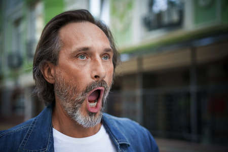 Handsome middle aged european man screaming on the street. Mature male in dressed up in casual jeans shirt stand with open mouth on street of old town. Yarning wide open moth man outdoors.の写真素材