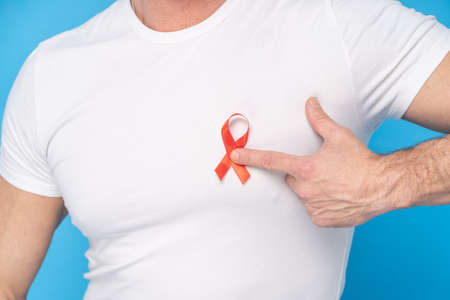 Man hold red ribbon bow AIDS awareness symbol on a heart wearing a white t-shirt isolated on a blue background. Modern medicine and healthcare. AIDS awareness concept.の写真素材
