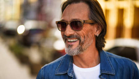 Middle aged handsome freelancer man with grey beard smiling looking sideways wearing casual denim shirt. Happy middle aged man in on streets of european city. Travel concept.の写真素材