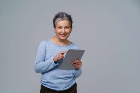 Smiling mature grey haired woman 50s holding digital tablet working or checking on social media. Pretty woman in 50s in blue blouse isolated on white. Older people and technologies. Toned image.の写真素材