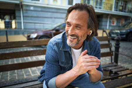 Handsome smiling mature man spend time talking on the phone or listening music using wireless earphones sitting on the bench in urban city, enjoying coffee wearing blue jeans shirt.の写真素材