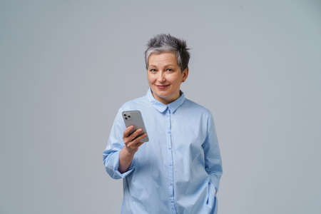 Gorgeous mature grey haired businesswoman hold smartphone in hand looking at camera working or shopping online or checking on social media. Pretty woman in blue blouse isolated on white background.の写真素材