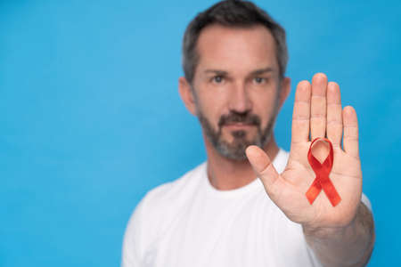 Man hold red ribbon bow AIDS awareness symbol on a heart wearing a white t-shirt isolated on a blue background. Modern medicine and healthcare. AIDS awareness concept.の写真素材