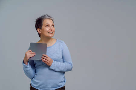 Dreaming beautiful mature grey haired woman in 50s holding digital tablet working or shopping online or checking on social media. Pretty woman in blue blouse isolated on white background. Toned image.の写真素材