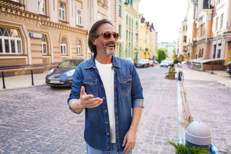 Portrait of a happy mature man outdoors in old town city smiling wearing jeans shirt and sunglasses. Happy mature man wearing sunglasses and looking sideways outdoor on a summer day.の写真素材
