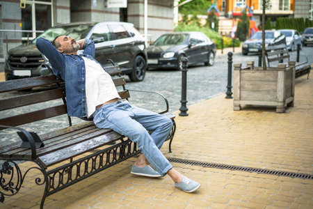 Relaxed handsome middle age man sits at the bench outdoors at urban city background enjoying free time with tablet standing next to him. Travel and people concept.の写真素材