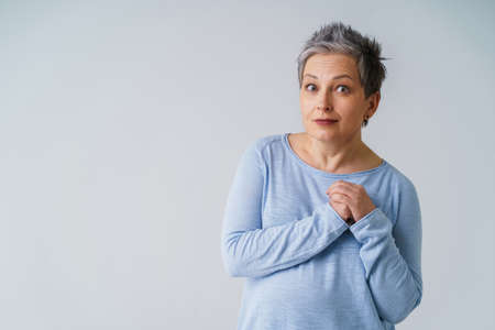 Amazed mature woman in 50s with grey hair posing with hands up next to heart and copy space on left isolated on white background. Copy space and place for product placement. Aged beauty. Toned image.の写真素材
