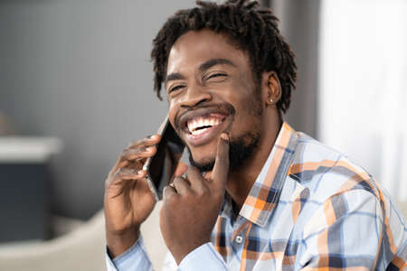 Happy smiling young African American man talking on the phone holding smartphone in hand looking away. Portrait of happy man having present conversation sitting on the sofa. Social media concept.の写真素材