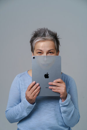 Mature grey haired woman hide hide behind apple ipad posting fake information or bad comments social media with drown evil face or alter ego on tablet isolated on white. Kyiv, Ukraine, May 13, 2022のeditorial素材