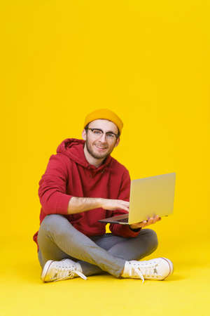 Happy young hipster man in red hoodie sitting on the floor holding laptop studying or working. Smart freelancer caucasian man with laptop isolated on yellow background.の写真素材