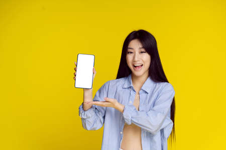 Young happy asian girl holding smartphone showing a white empty screen and exciting to win isolated on yellow background. Great offer. Product placement. Mobile App Advertisement.の写真素材