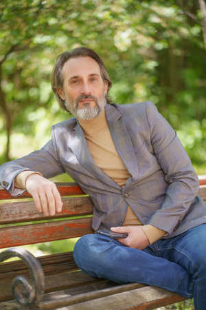Mature handsome freelancer man with grey beard, looking thoughtful at the camera sitting on the bench at the park wearing casual jacket and jeans holding smartphone in hand.の写真素材