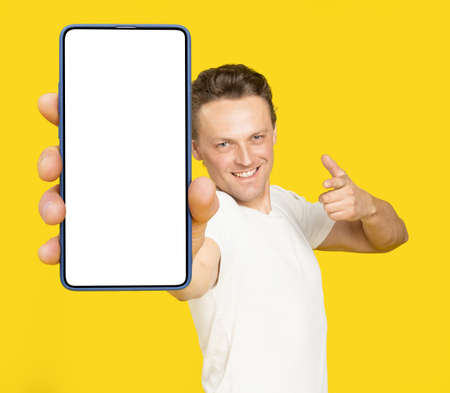 Holding huge smartphone with white screen handsome blond man pointing at white empty screen, wearing white t-shirt and jeans isolated on yellow background. Mobile app advertisement, great offer.の写真素材