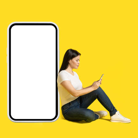 Sitting on the floor middle aged asian woman with phone in hands leaned on giant, huge smartphone with white screen wearing white t-shirt and jeans isolated on yellow background. Free space mock up.の写真素材