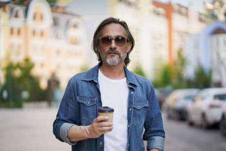 Handsome middle aged man with grey beard walking in the city holding take away coffee in paper cup. Handsome man traveling enjoying free time. Creative man on a coffee break outdoors.の写真素材