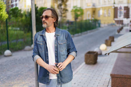 Emotionally exhausted middle age crisis mature man leaned on road sign holding paper cup off coffee. Experiencing hangover middle age alcoholic man drink using paper cup to hide alcohol.の写真素材