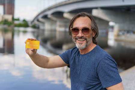 Middle aged man showing debit, credit card sitting outdoors next to river working using digital...