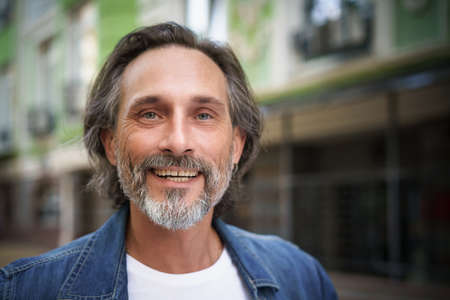 Happy handsome middle aged man standing outdoors on street of city looking at camera with a charming smile. Middle aged grey bearded man traveling in city smiling on camera wearing casual.の写真素材