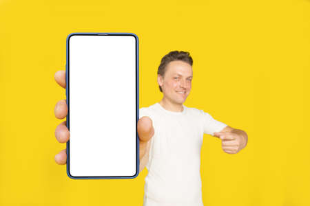 Huge smartphone in hand of handsome blond man pointing at white empty screen, wearing white t-shirt and jeans isolated on yellow background. Mobile app advertisement, great offer.の写真素材