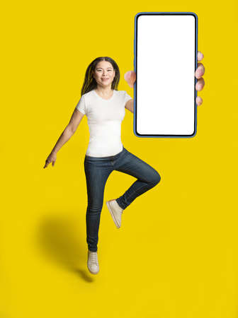 Middle aged asian woman freezed jumping with white screen smartphone wearing casual outfit, isolated on yellow background, mobile app advertisement. Full length portrait of joyful asian woman.の写真素材