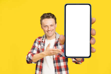 Introducing new app happy holding giant, huge smartphone with white screen blond man, wearing red plaid shirt. Man with phone display mock up isolated on yellow background. Mobile app advertisement.の写真素材