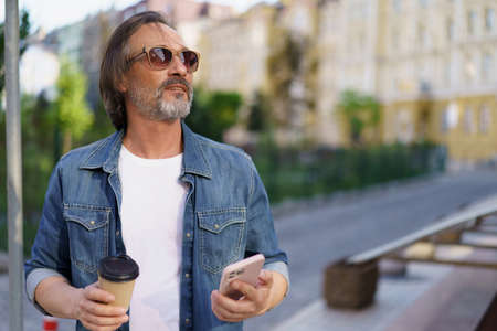 Middle aged man got distracted from mobile phone enjoying coffee outdoors looking at sunset. Middle aged man looking away at street while enjoying free time off work. Business on the go.の写真素材