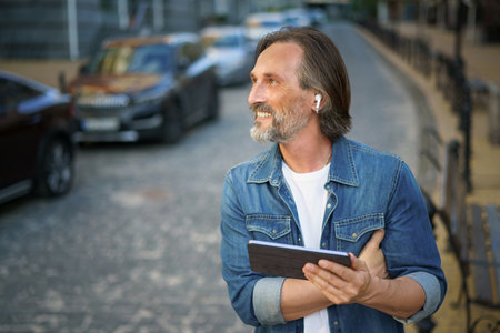 Middle aged freelancer man use gps or calling digital tablet while walking on urban streets. Mature man use wireless earphones talking or listening music while travel in old town streets.の写真素材