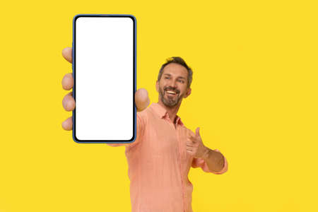 Middle aged grey haired man pointing at smartphone in hand amazed smiling looking at smartphone wearing peach shirt isolated on yellow. Mature fit man with phone, mobile app advertisement.の写真素材